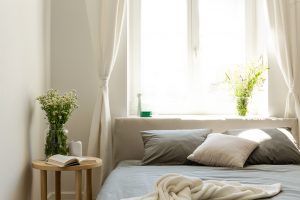 Sunny natural style bedroom with a bed in the middle, gray linen and pillows, and a vanilla blanket. A lamp and a stool with fresh flowers next to the bed and a big window behind, looking out at the Indianapolis skyline after couples therapy